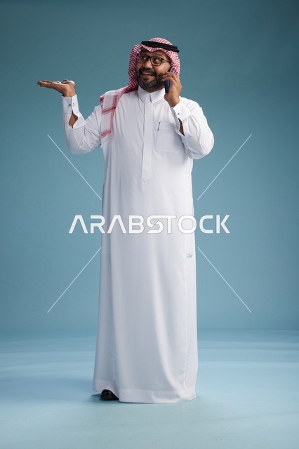 بورتريه لرجل سعودي يرتدي الثوب الابيض والشماغ يتحدث بالهاتف المحمول بإيماءة تعبيرية , الجسم بالكامل, خلفيه زرقاء