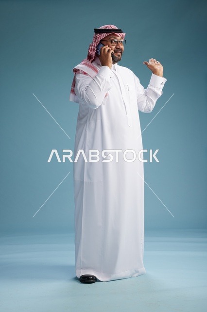 بورتريه لرجل سعودي يرتدي الثوب الابيض والشماغ يتحدث بالهاتف المحمول بإيماءة تعبيرية , الجسم بالكامل, خلفيه زرقاء