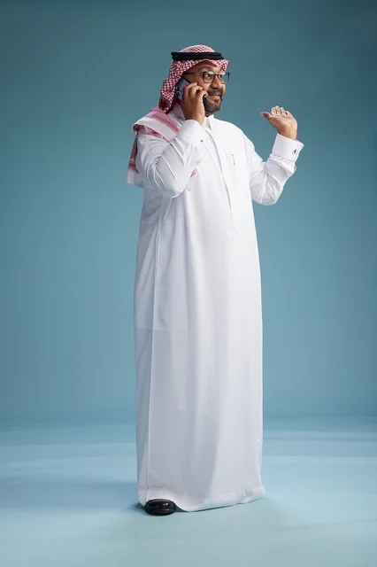 A portrait of a Saudi man wearing a white thobe and a shemagh, talking on a mobile phone with an expressive gesture, full body, blue background.