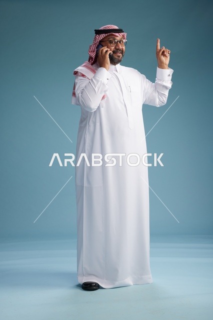 بورتريه لرجل سعودي يرتدي الثوب الابيض والشماغ يتحدث بالهاتف المحمول بإيماءة تعبيرية , الجسم بالكامل, خلفيه زرقاء