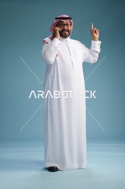 بورتريه لرجل سعودي يرتدي الثوب الابيض والشماغ يتحدث بالهاتف المحمول بإيماءة تعبيرية , الجسم بالكامل, خلفيه زرقاء