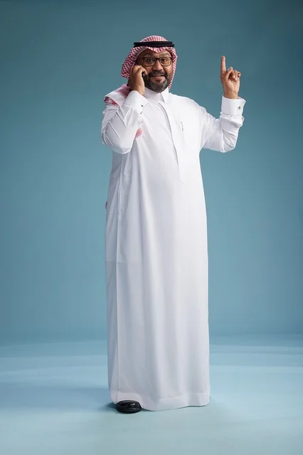 A portrait of a Saudi man wearing a white thobe and a shemagh, talking on a mobile phone with an expressive gesture, full body, blue background.
