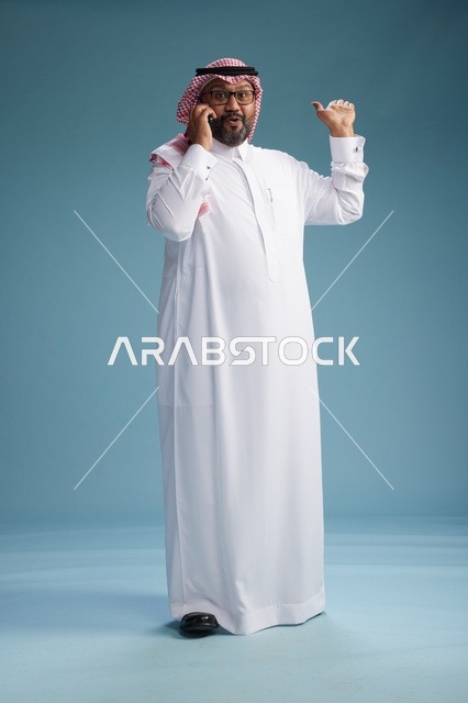 بورتريه لرجل سعودي يرتدي الثوب الابيض والشماغ يتحدث بالهاتف المحمول بإيماءة تعبيرية , الجسم بالكامل, خلفيه زرقاء