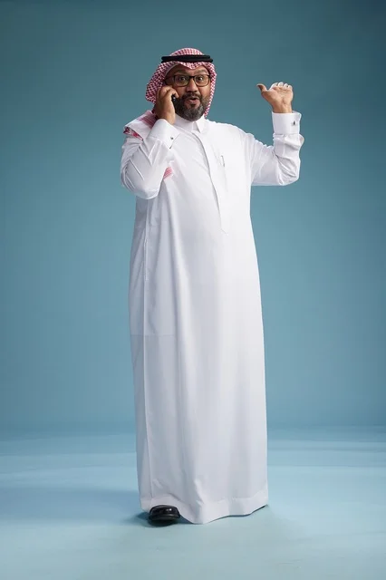 A portrait of a Saudi man wearing a white thobe and a shemagh, talking on a mobile phone with an expressive gesture, full body, blue background.