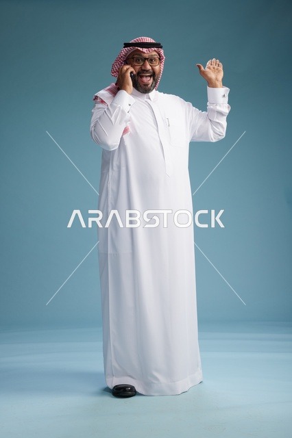 بورتريه لرجل سعودي يرتدي الثوب الابيض والشماغ يتحدث بالهاتف المحمول بإيماءة تعبيرية , الجسم بالكامل, خلفيه زرقاء