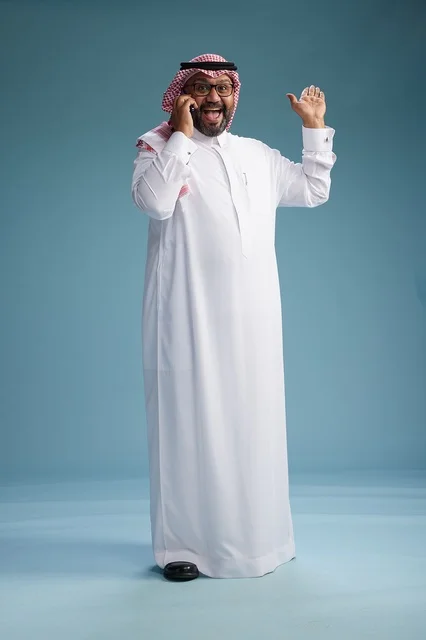 A portrait of a Saudi man wearing a white thobe and a shemagh, talking on a mobile phone with an expressive gesture, full body, blue background.