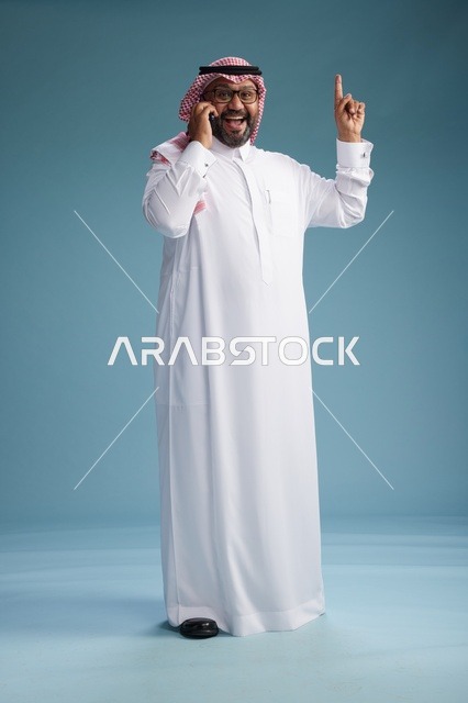 بورتريه لرجل سعودي يرتدي الثوب الابيض والشماغ يتحدث بالهاتف المحمول بإيماءة تعبيرية , الجسم بالكامل, خلفيه زرقاء