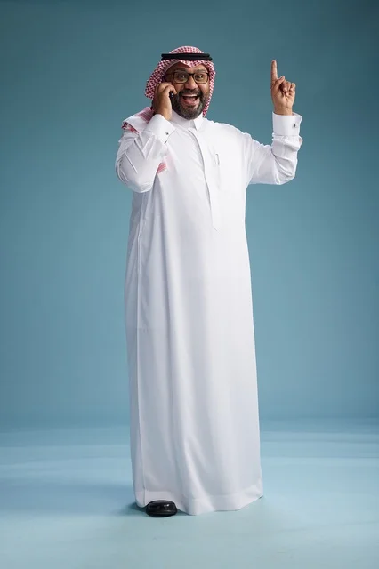 A portrait of a Saudi man wearing a white thobe and a shemagh, talking on a mobile phone with an expressive gesture, full body, blue background.