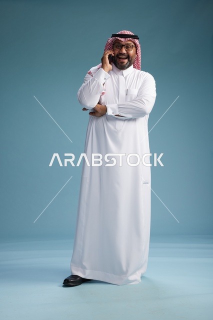بورتريه لرجل سعودي يرتدي الثوب الابيض والشماغ يتحدث بالهاتف المحمول بإيماءة تعبيرية , الجسم بالكامل, خلفيه زرقاء