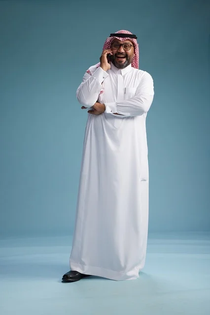 A portrait of a Saudi man wearing a white thobe and a shemagh, talking on a mobile phone with an expressive gesture, full body, blue background.
