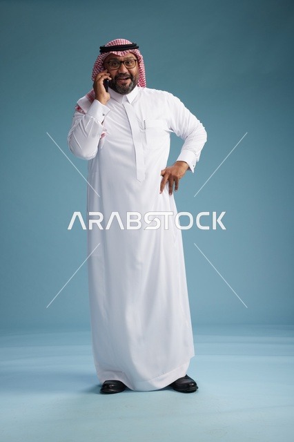 بورتريه لرجل سعودي يرتدي الثوب الابيض والشماغ يتحدث بالهاتف المحمول بإيماءة تعبيرية , الجسم بالكامل, خلفيه زرقاء
