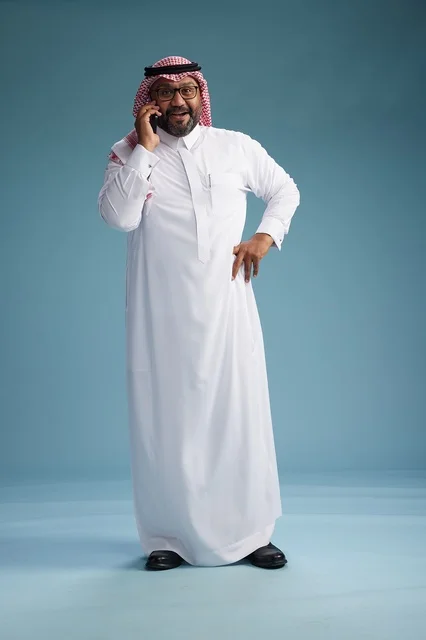 A portrait of a Saudi man wearing a white thobe and a shemagh, talking on a mobile phone with an expressive gesture, full body, blue background.
