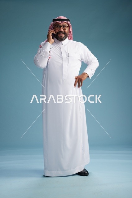 A portrait of a Saudi man wearing a white thobe and a shemagh, talking on a mobile phone with an expressive gesture, full body, blue background.