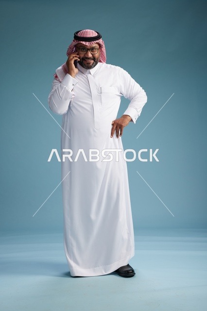 بورتريه لرجل سعودي يرتدي الثوب الابيض والشماغ يتحدث بالهاتف المحمول بإيماءة تعبيرية , الجسم بالكامل, خلفيه زرقاء