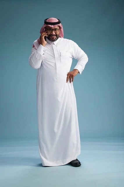 A portrait of a Saudi man wearing a white thobe and a shemagh, talking on a mobile phone with an expressive gesture, full body, blue background.