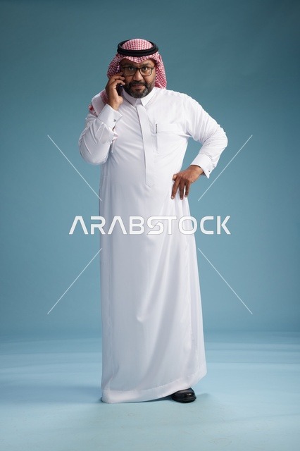 بورتريه لرجل سعودي يرتدي الثوب الابيض والشماغ يتحدث بالهاتف المحمول بإيماءة تعبيرية , الجسم بالكامل, خلفيه زرقاء