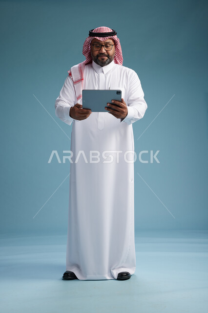 رجل سعودي بالزي التقليدي واقف يستخدم جهاز لوحي, استخدام جهاز التابليت, خلفيه زرقاء, كامل الجسم