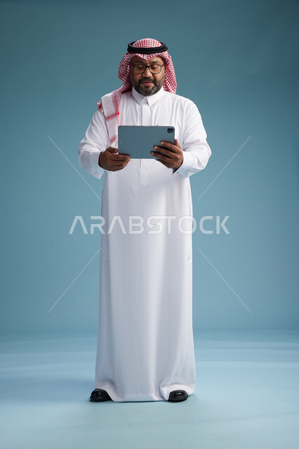 رجل سعودي بالزي التقليدي واقف يستخدم جهاز لوحي, استخدام جهاز التابليت, خلفيه زرقاء, كامل الجسم