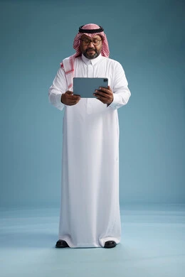 A Saudi man in traditional attire standing using a tablet, using the tablet, blue background, full body.
