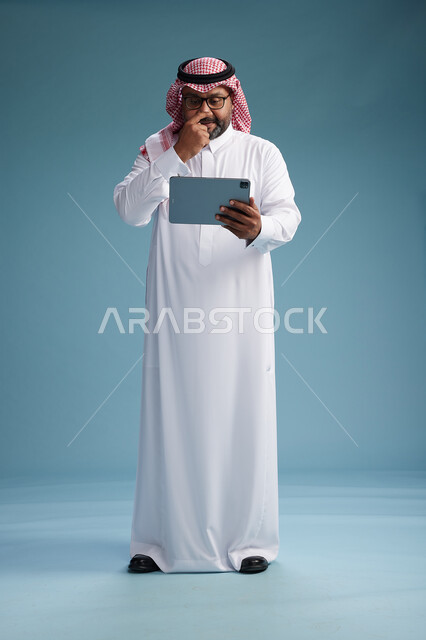 A Saudi man in traditional attire standing using a tablet, using the tablet, blue background, full body.
