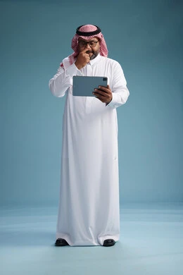 A Saudi man in traditional attire standing using a tablet, using the tablet, blue background, full body.