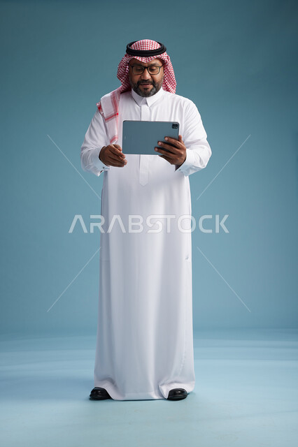 رجل سعودي بالزي التقليدي واقف يستخدم جهاز لوحي, استخدام جهاز التابليت, خلفيه زرقاء, كامل الجسم