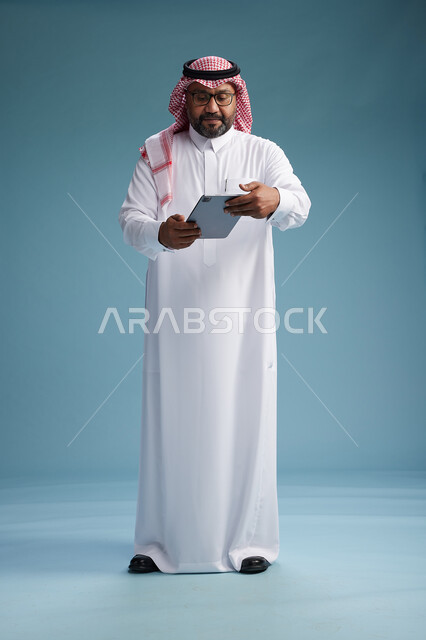 A Saudi man in traditional attire standing using a tablet, using the tablet, blue background, full body.