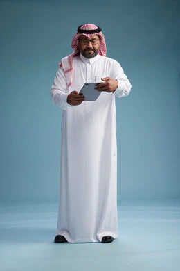 A Saudi man in traditional attire standing using a tablet, using the tablet, blue background, full body.