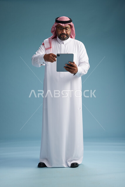 A Saudi man in traditional attire standing using a tablet, using the tablet, blue background, full body.