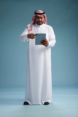 A Saudi man in traditional attire standing using a tablet, using the tablet, blue background, full body.