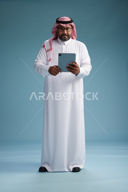 A Saudi man in traditional attire standing using a tablet, using the tablet, blue background, full body.