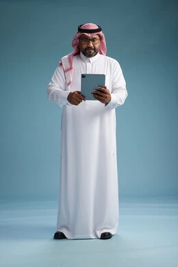 A Saudi man in traditional attire standing using a tablet, using the tablet, blue background, full body.