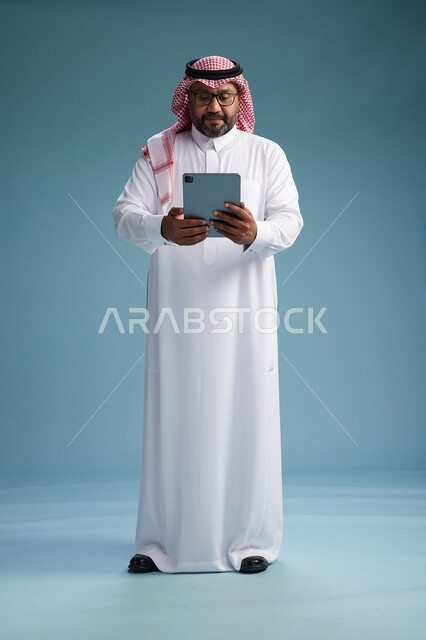 A Saudi man in traditional attire standing using a tablet, using the tablet, blue background, full body.