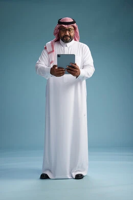 A Saudi man in traditional attire standing using a tablet, using the tablet, blue background, full body.