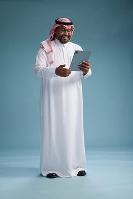 A Saudi man in traditional attire standing using a tablet, using the tablet, blue background, full body.