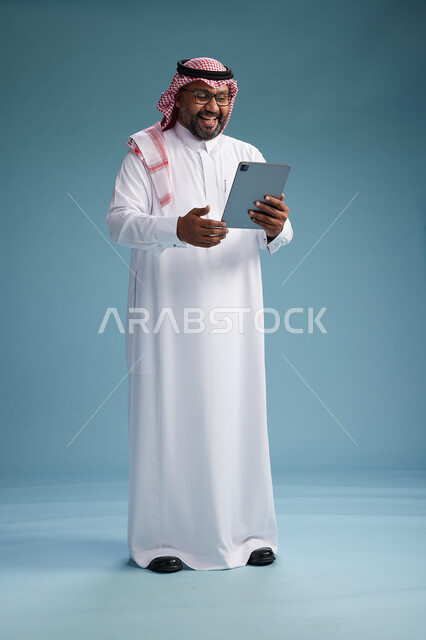 رجل سعودي بالزي التقليدي واقف يستخدم جهاز لوحي, استخدام جهاز التابليت, خلفيه زرقاء, كامل الجسم