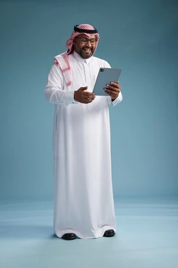 رجل سعودي بالزي التقليدي واقف يستخدم جهاز لوحي, استخدام جهاز التابليت, خلفيه زرقاء, كامل الجسم