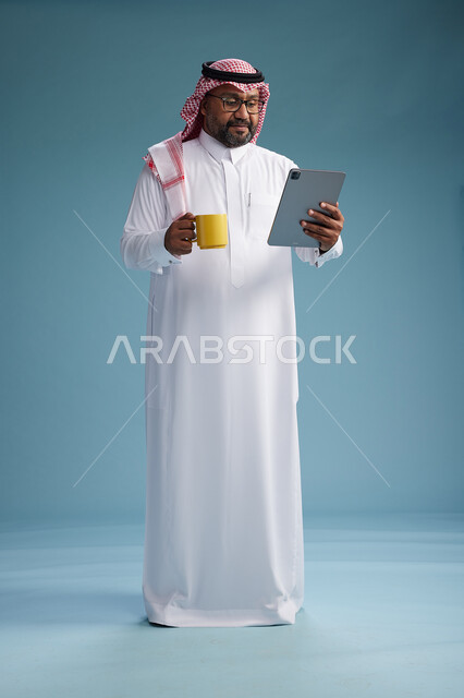 بورتريه لرجل عربي  سعودي خليجي بالزي التقليدي واقف يستخدم جهاز لوحي  ويمسك كوب اصفر ,الاستمتاع بمشروب ساخن, الجسم بالكامل, خلفيه زرقاء