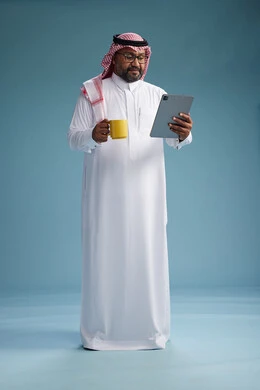 A portrait of an Arab Saudi Gulf man in traditional attire standing while using a tablet and holding a yellow cup, enjoying a hot drink, full body, blue background.