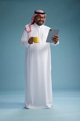 A portrait of an Arab Saudi Gulf man in traditional attire standing while using a tablet and holding a yellow cup, enjoying a hot drink, full body, blue background.