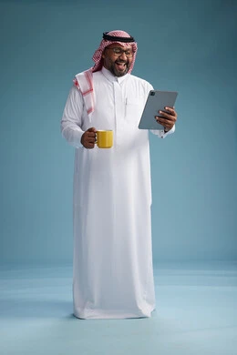 A portrait of an Arab Saudi Gulf man in traditional attire standing while using a tablet and holding a yellow cup, enjoying a hot drink, full body, blue background.