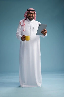 A portrait of an Arab Saudi Gulf man in traditional attire standing while using a tablet and holding a yellow cup, enjoying a hot drink, full body, blue background.