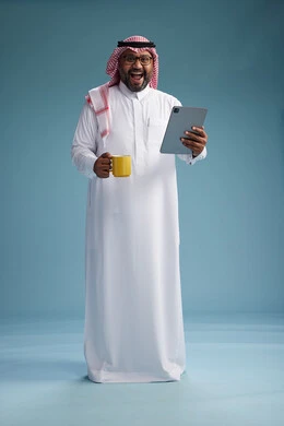 بورتريه لرجل عربي  سعودي خليجي بالزي التقليدي واقف يستخدم جهاز لوحي  ويمسك كوب اصفر ,الاستمتاع بمشروب ساخن, الجسم بالكامل, خلفيه زرقاء