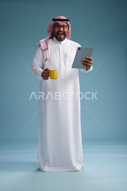 بورتريه لرجل عربي  سعودي خليجي بالزي التقليدي واقف يستخدم جهاز لوحي  ويمسك كوب اصفر ,الاستمتاع بمشروب ساخن, الجسم بالكامل, خلفيه زرقاء