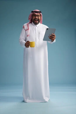 A portrait of an Arab Saudi Gulf man in traditional attire standing while using a tablet and holding a yellow cup, enjoying a hot drink, full body, blue background.