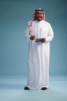 A Saudi man in traditional attire standing using a tablet, using the tablet, blue background, full body.