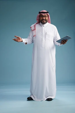 A Saudi man in traditional attire standing using a tablet, using the tablet, blue background, full body.