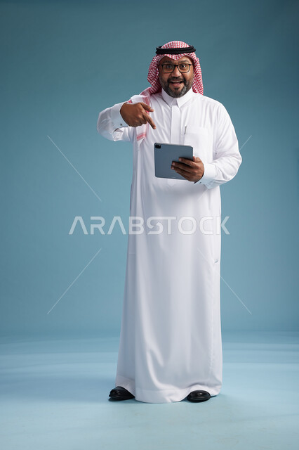 A Saudi man in traditional attire standing using a tablet, using the tablet, blue background, full body.