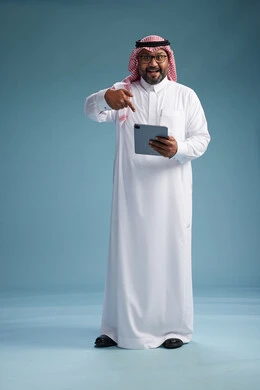 A Saudi man in traditional attire standing using a tablet, using the tablet, blue background, full body.