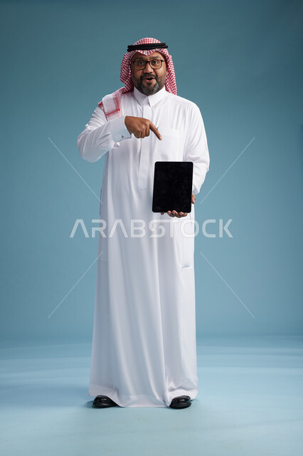 A Saudi man in traditional attire standing using a tablet, using the tablet, blue background, full body.